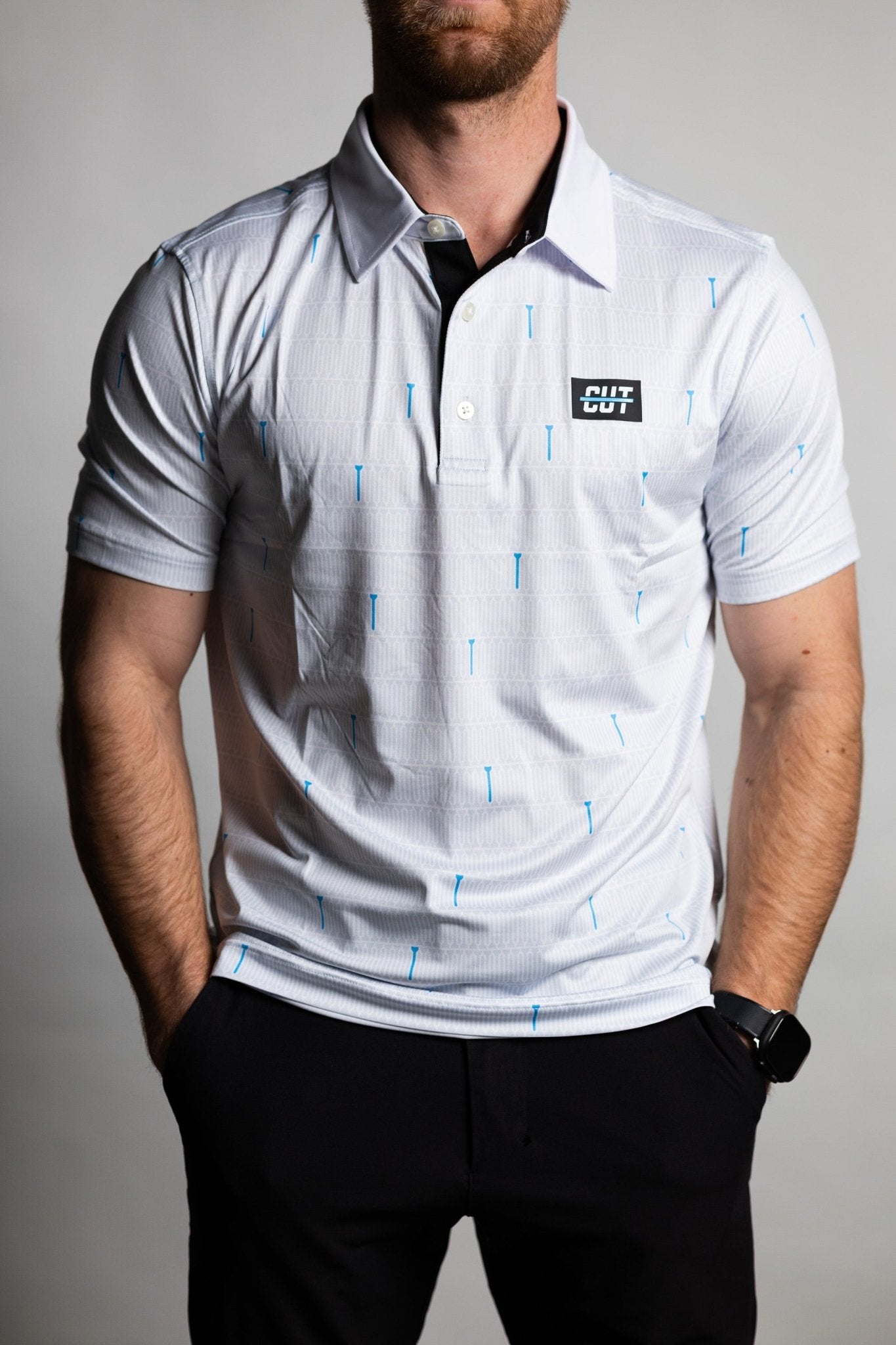 Man wearing a white Teal "Tee" Polo with blue golf tee pattern and black pants, hands in pockets