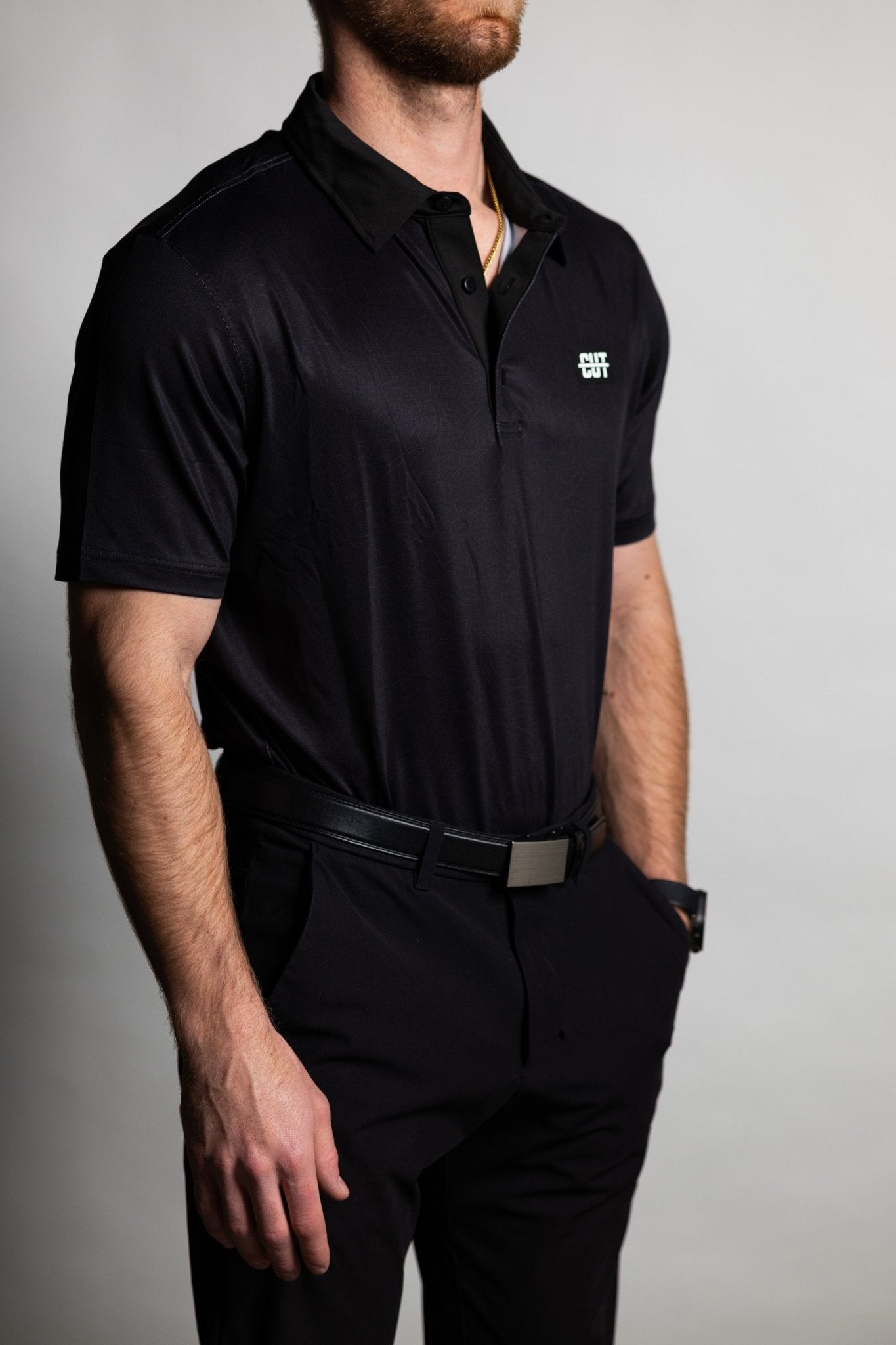 Man wearing a sleek Deep Topo Black Polo with black pants and a belt, styled for golf apparel and mens polos.