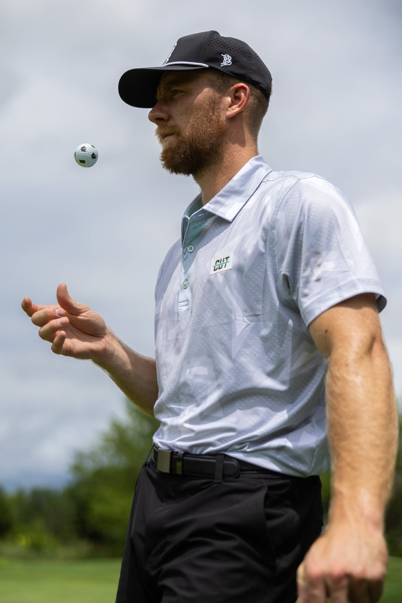 Man wearing Future Grey Pin Polo tossing a golf ball outdoors on a sunny day with black cap and pants