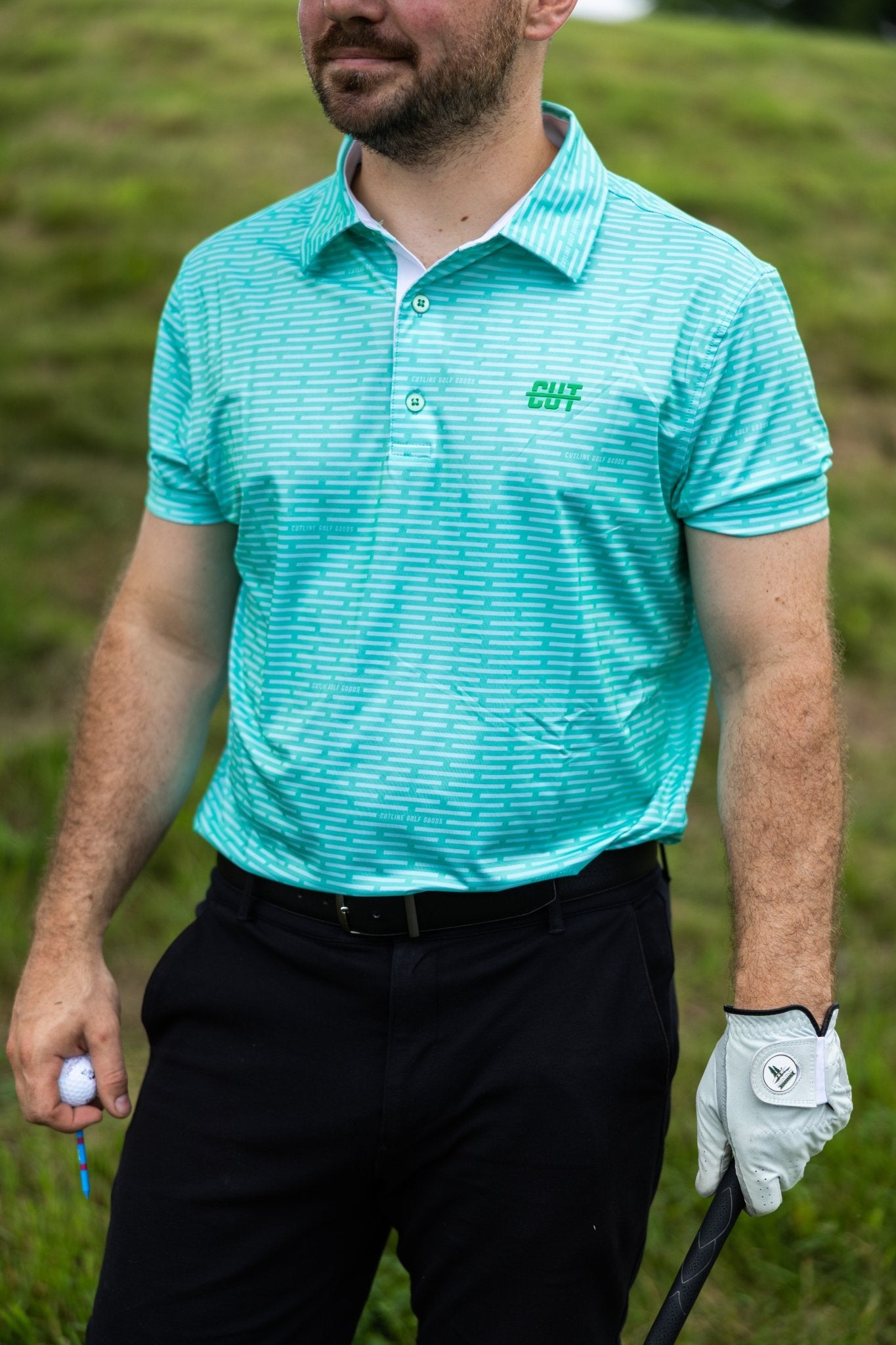 Man wearing a turquoise Minted Lines Polo shirt holding a golf ball and club on a grassy course