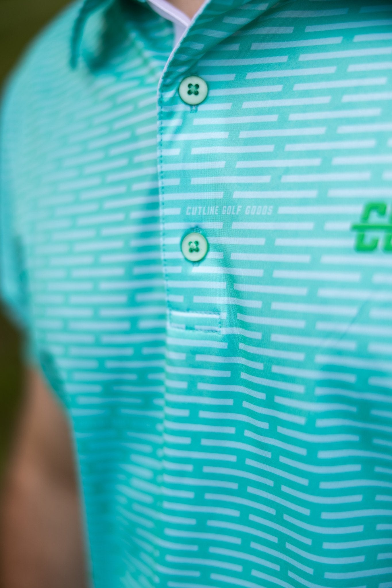 Close-up of a turquoise Minted Lines Polo with horizontal white line pattern and green buttons on golf apparel