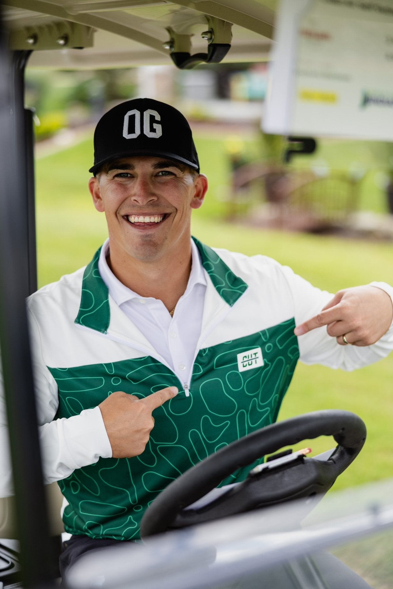 Man smiling in golf cart wearing Green Topo Athletic Q-Zip and a black OG cap on a sunny day