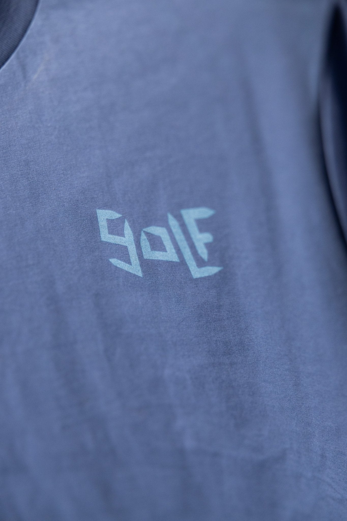 Close-up of blue Play More Golf T-Shirt with abstract golf text design in light blue on soft fabric