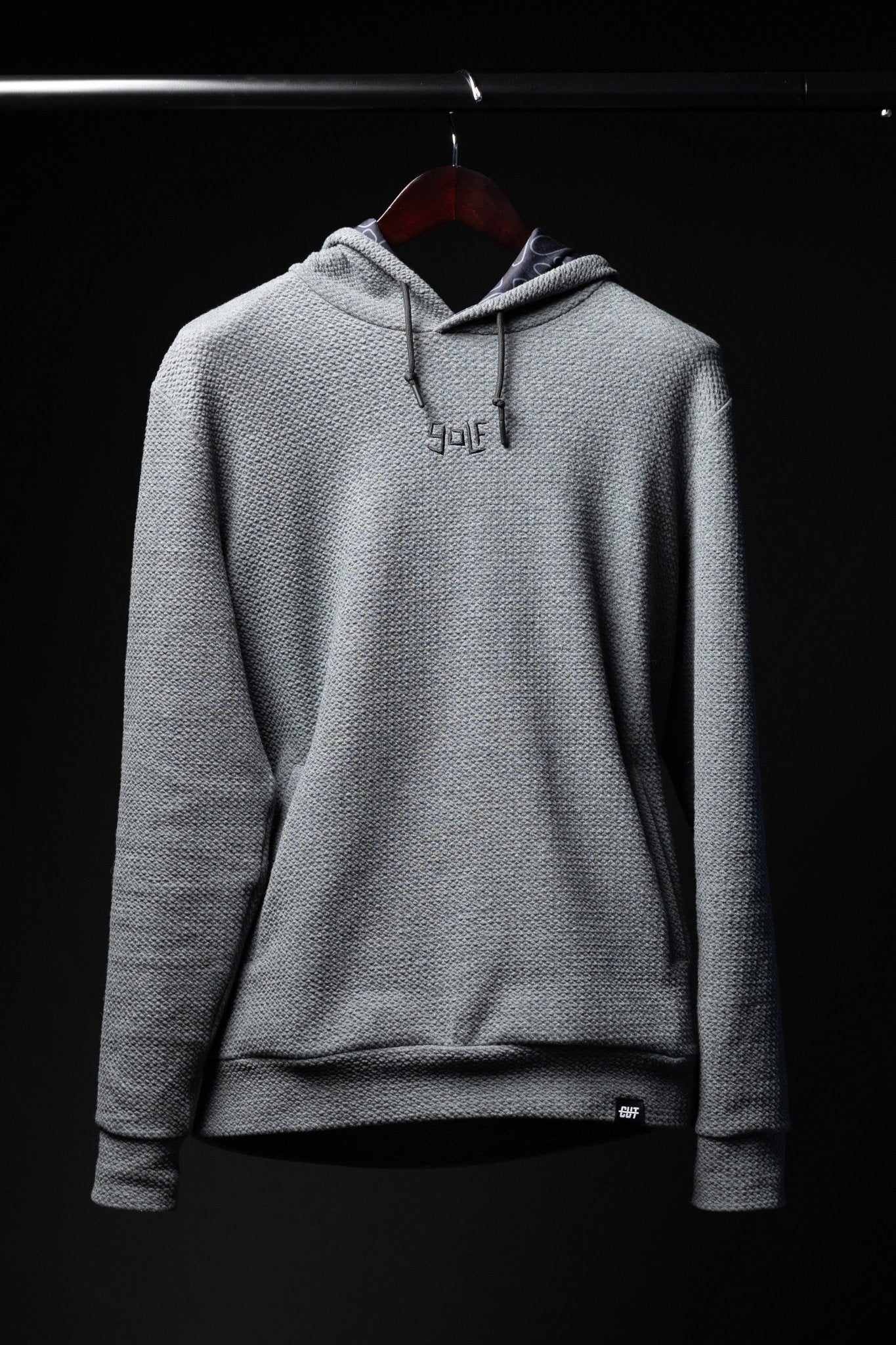 Waffle Knit Cobalt Grey GOLF Hoodie hanging on wooden hanger against black background in soft lighting