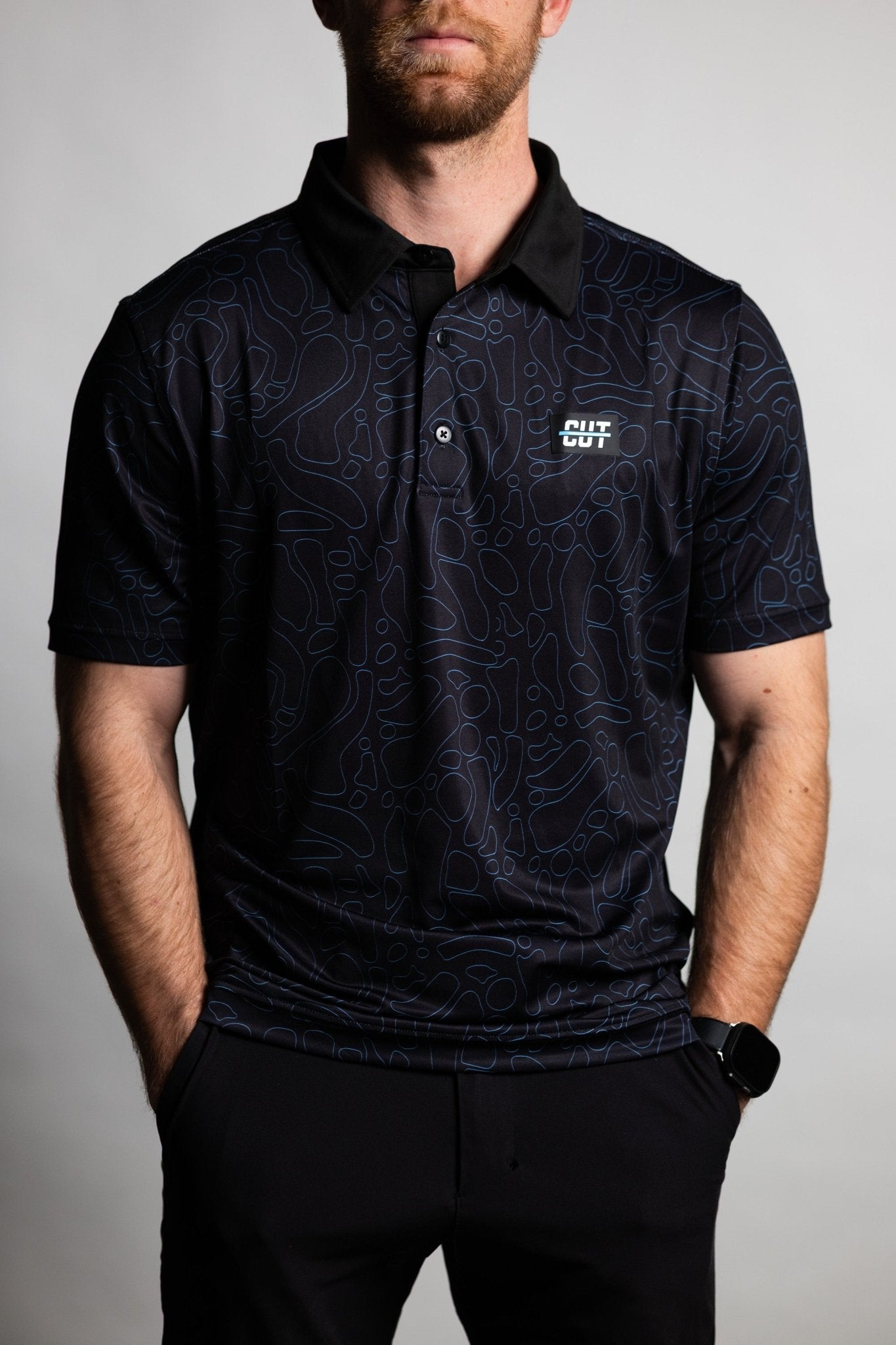 Man wearing a Midnight Teal Topo Polo with dark teal contour pattern and black collar, hands in pockets.