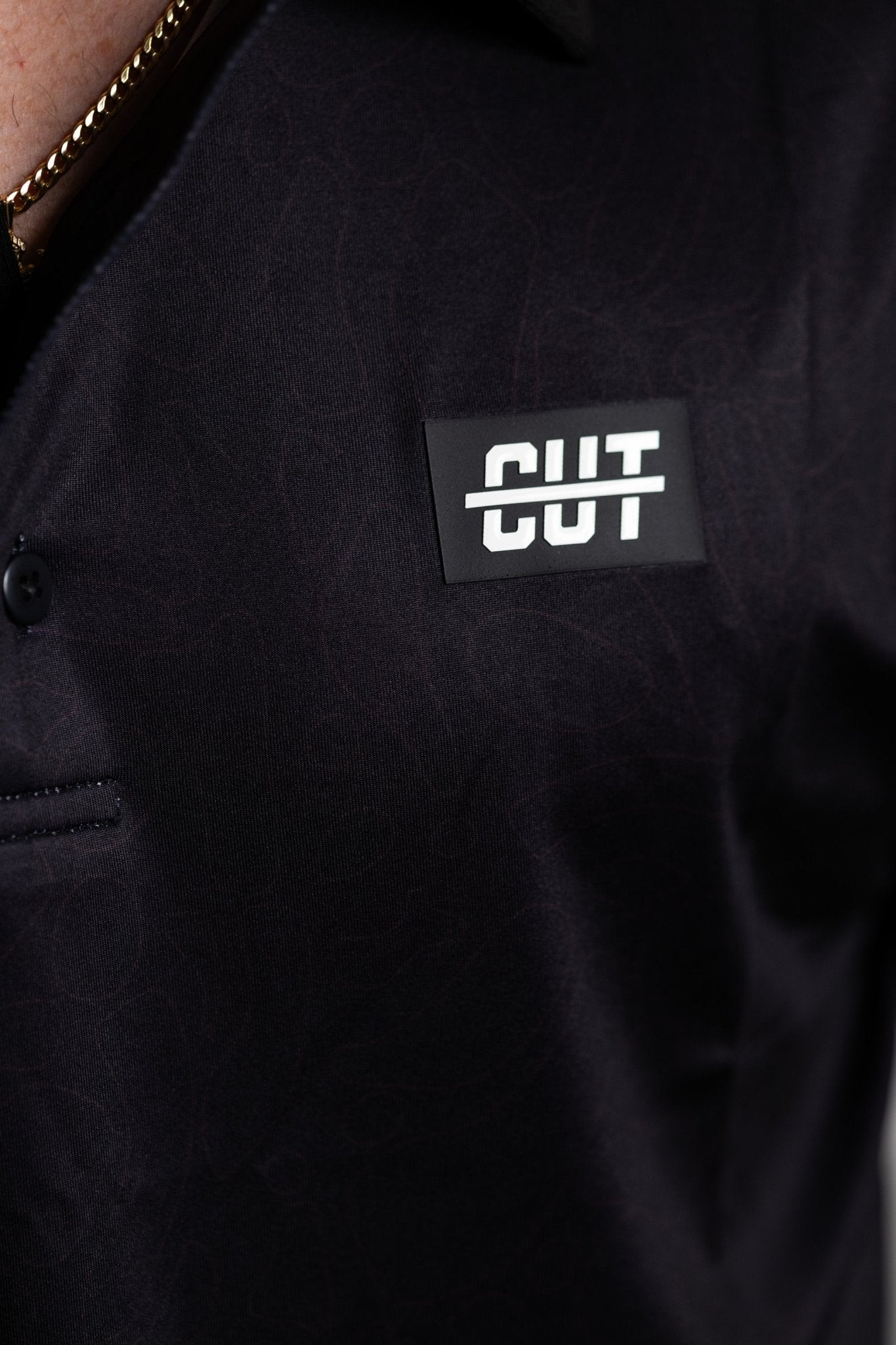 Close-up of the Deep Topo Black Polo showing subtle topographic pattern and a Cutline Golf Goods logo patch on chest.