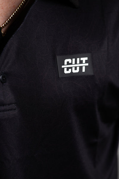 Close-up of the Deep Topo Black Polo showing subtle topographic pattern and a Cutline Golf Goods logo patch on chest.