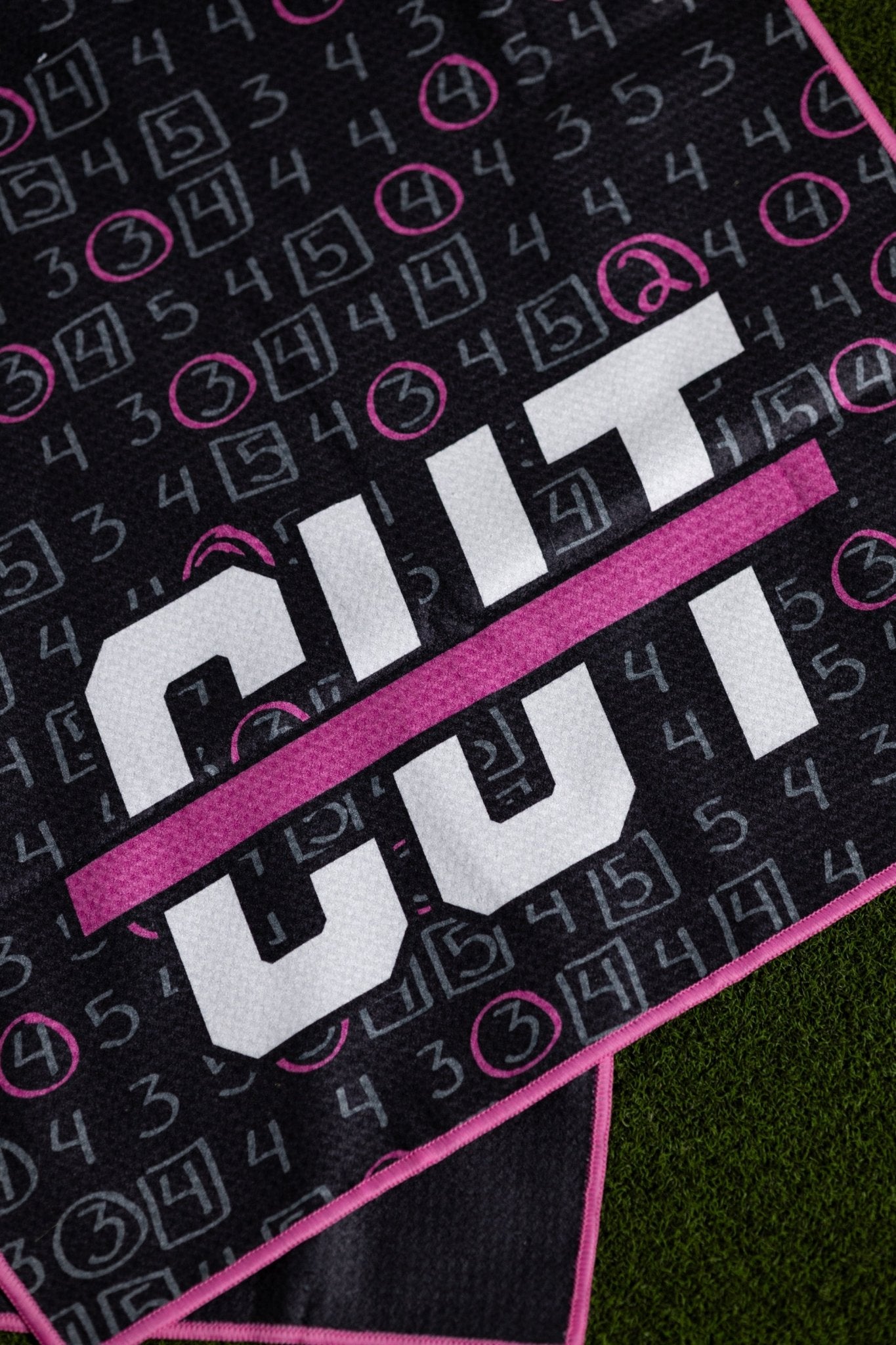 Close-up of a black Pink Scorecard Oversized Towel with white and pink scorecard design and pink trim on grass