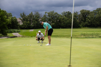 Man wearing Minted Lines Polo preparing to putt on golf green with another man watching on a cloudy day