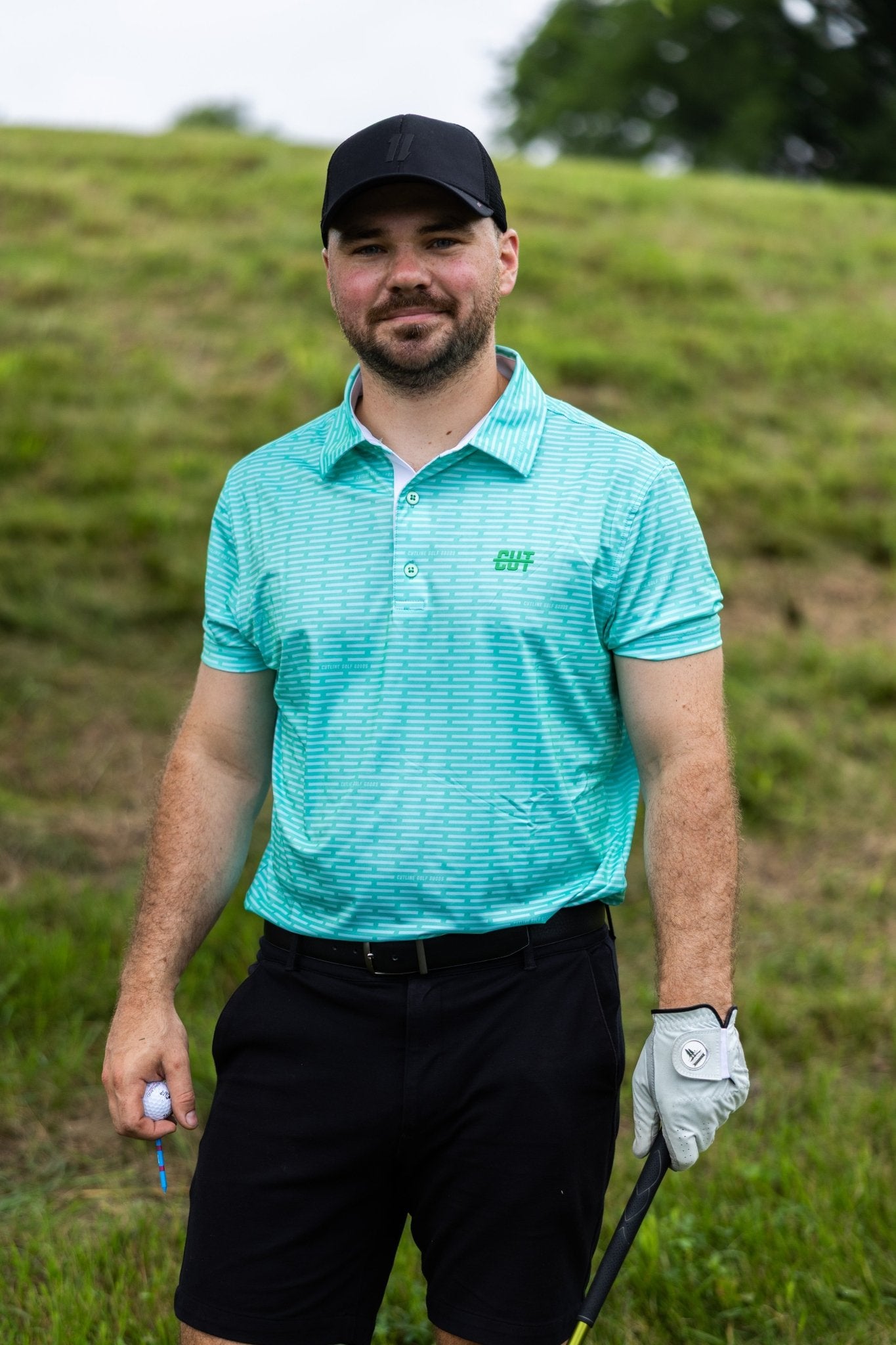 Man wearing a turquoise Minted Lines Polo shirt and black shorts on a golf course holding a golf club and ball