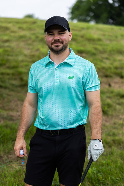 Man wearing a turquoise Minted Lines Polo shirt and black shorts on a golf course holding a golf club and ball