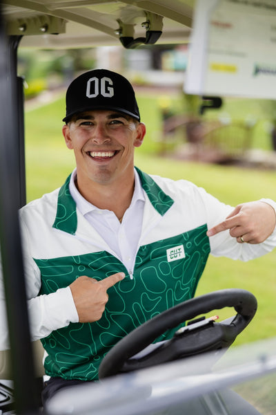 Man smiling in golf cart wearing Green Topo Athletic Q-Zip and a black OG cap on a sunny day