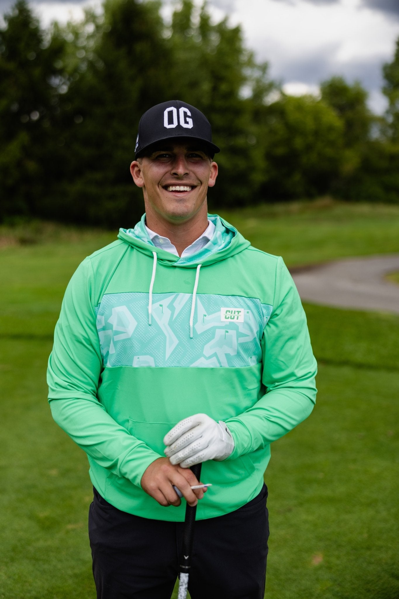 Man smiling on golf course wearing a Mint Future Tech Athletic Hoodie and a black OG hat holding a golf club