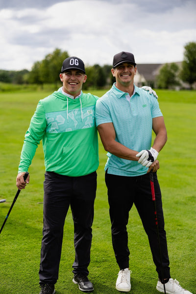 Two men smiling on a golf course, one wearing a Minted Lines Polo and golf gloves, holding a golf club.