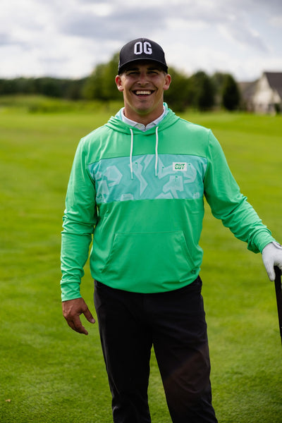 Man smiling on golf course wearing Mint Future Tech Athletic Hoodie and black cap holding golf club