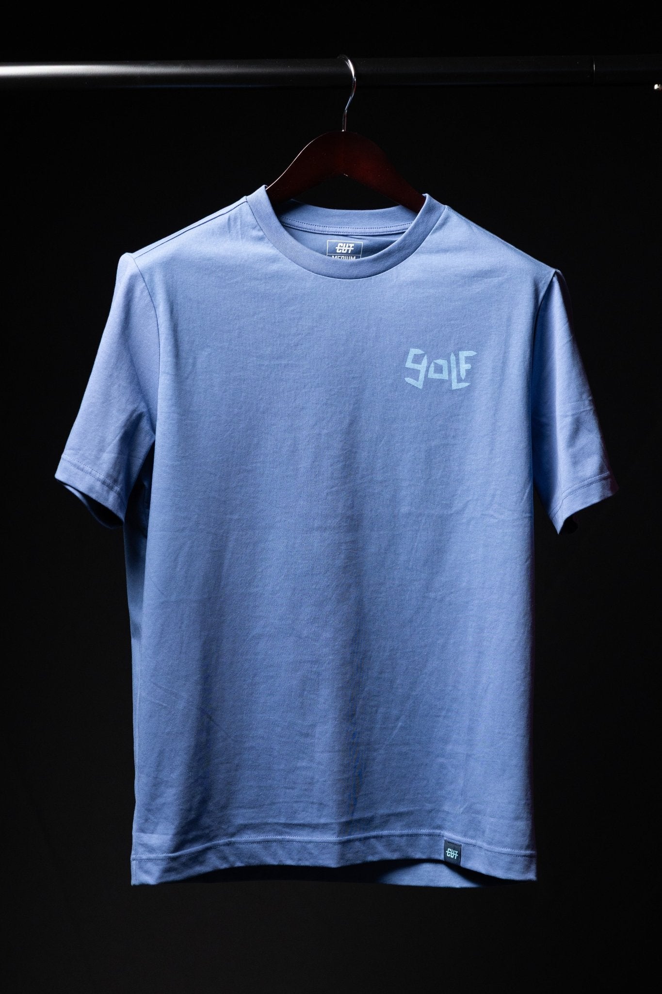 Light blue Play More Golf T-Shirt with subtle golf graphic on chest hanging against black background